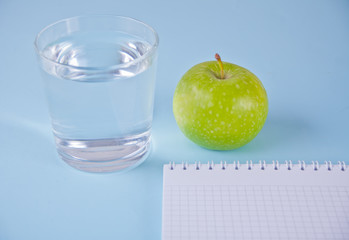 Fresh apple, glass of water and notebook on blue background.