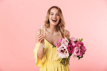 Beautiful amazing young blonde woman posing isolated over pink wall background holding flowers drinking champagne.