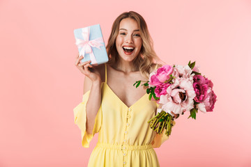 Young blonde woman posing isolated over pink wall background holding flowers holding present gift box.