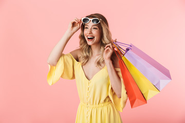 Beautiful shocked young blonde woman posing isolated over pink wall background holding shopping bags.