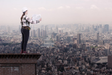 Female engineer holding construction plans standing on the roof top above a city