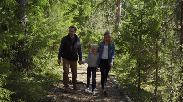 Happy family of father, mother and daughter walking along path in coniferous forest. Family talking cheerfully and showing nature to little girl