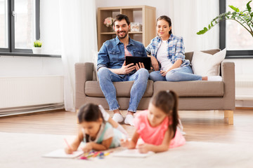 family, leisure and people concept - happy mother and father with tablet pc computer sitting on sofa and looking at daughters drawing at home