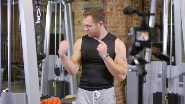 Male blogger demonstrating his biceps, speaking on camera and showing how to do cable pushdown in gym while recording video lesson for vlog