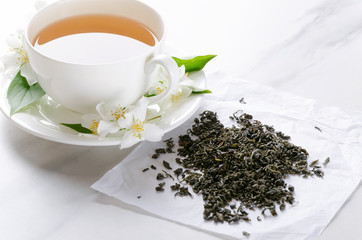 Cup of hot green tea with jasmine flavor, fresh jasmine flowers and dry green tea on the paper, marble kitchen table