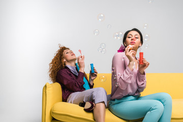 redhead girl and african american woman blowing soap bubbles while sitting on yellow sofa on white