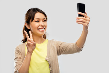 technology and people concept - happy asian woman taking selfie by smartphone and showing peace over grey background