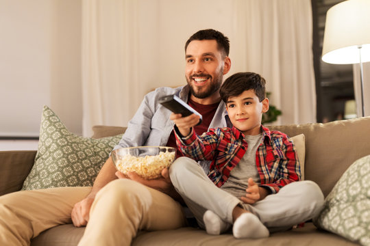 family, childhood, fatherhood, technology and people concept - happy father and little son with popcorn and remote control watching tv at home in evening