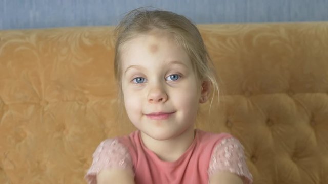 Happy funny little blue-eyed girl with blond hair sends a kiss sits on yellow sofa at home