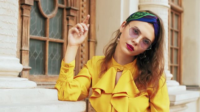 Street fashion portrait of young beautiful elegant girl wearing trendy purple color sunglasses, headband, stylish yellow blouse with jabot, posing in old european city