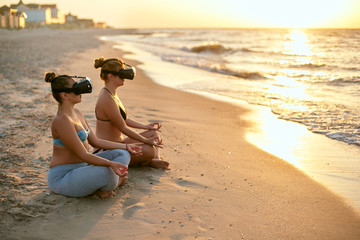 Women use VR glasses for deeper immersion. Two females doing group yoga meditation on the beach in...