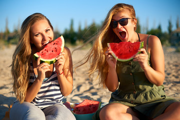 Happy young woman in glasses eating watermelon on the sandy beach on vacation. Girl joyfully holding fresh watermelon skip in her hands and have fun with hair fluttering in the wind. Youth lifestyle.