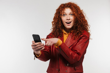 Naklejka premium Image of excited redhead woman 20s wearing leather jacket smiling and holding smartphone