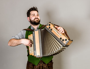 cool young musician with black beard and leather trousers and traditional costume and accordion is posing in front of grey background