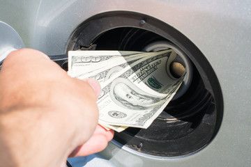 Male hand puts a band of US dollars in a fuel tank of a silver car, symbolizing high cost of fuel