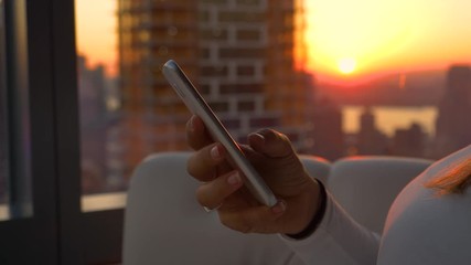 CLOSE UP, DOF: Sunset illuminates the stunning cityscape surrounding the unrecognizable girl texting from her hotel room high above New York City. Young woman browses the internet on her cell phone - Powered by Adobe