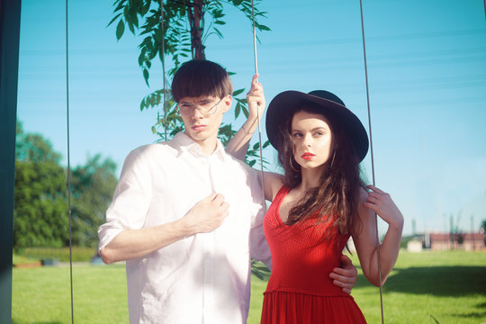 Beautiful fashionable couple against the sky and the beach . Fashionable hipsters. Girl in a red dress and black big hat. A man with glasses and a white shirt. Model pouse at holiday, relaxation.