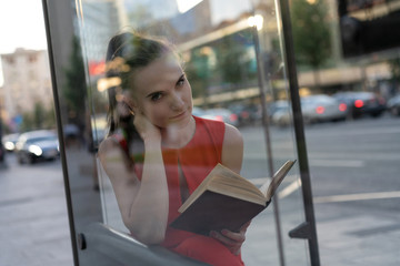 Young beautiful european girl with a book in her hands sitting at the bus stop. The view through the glass, a pleasant evening atmosphere of tranquility. Copy space. Lifestyle