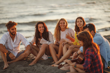 Happy friends sitting on the beach singing and playing guitar during the sunset