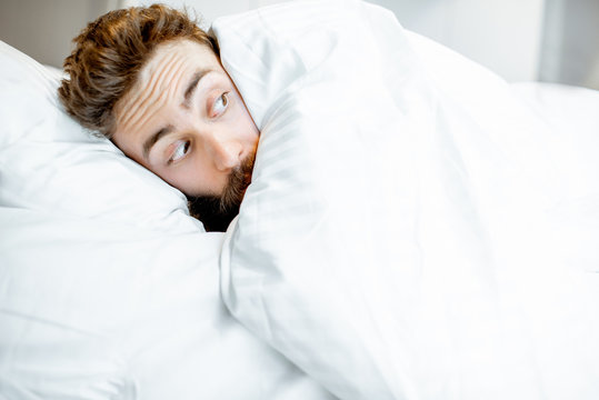 Man hiding in bed under the white sheets feeling scared alone