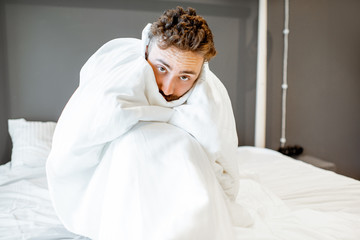Frustrated man with emotional insanity sitting on the bed covered with white sheets. Concept of insomnia or emotional problems