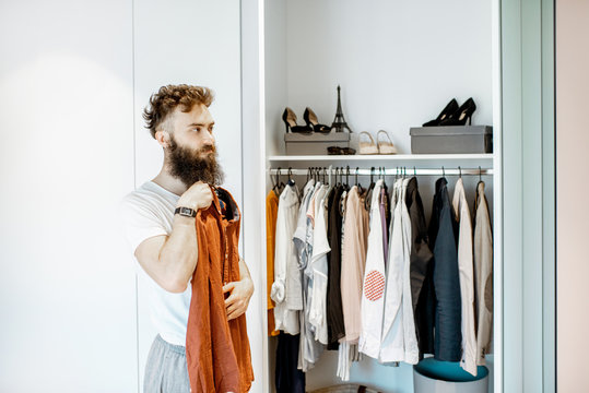 Bearded man trying shirt, choosing clothes to wear in the wardrobe at home