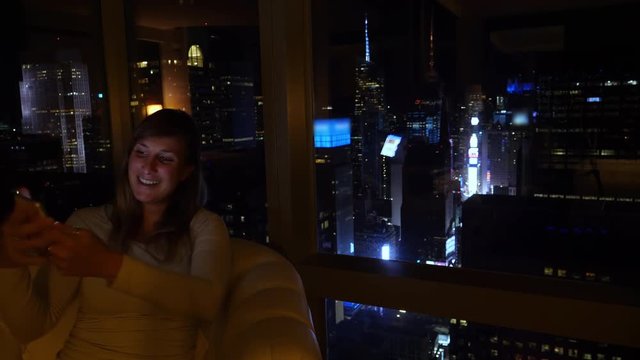 CLOSE UP: Girl points at Times Square while video calling friends at night. Young Caucasian woman skypes with her family from a luxury hotel room with a spectacular view of the New York's cityscape.