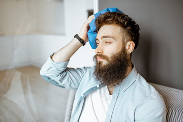 Bearded man with headache applying ice bag while sitting on the couch at home