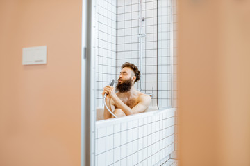 Joyful bearded man washing in the bathtub, having fun singing into the shower in the bathroom, view...