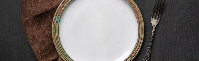 top view of vintage white empty round plate on black wooden table near brown napkin and silver fork, panoramic shot