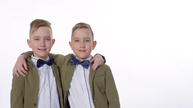 Waist-up portrait shot of 10-year-old Caucasian identical twin brothers, smartly dressed in blazers, white shirts and bowties, hugging and posing together for photos in studio against white background