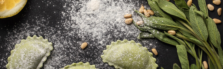 panoramic shot of green raw ravioli, pine nuts, herb and flour on black background