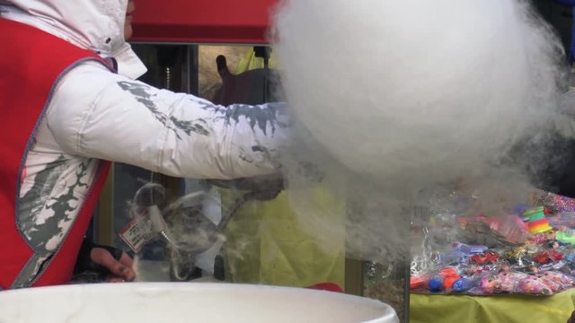 A person spins big white cotton candy with an automated machine at a funfair. A seller makes white candy floss with a modern machine at the amusement park.