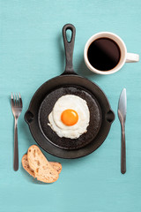 black frying pan with scrambled eggs, fork, knife, with cup of coffee two pieces of bread on a turquoise tablecloth