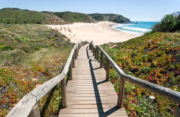 Wooden steps to the sunny beach in Portugal town. Ocean waters and green hills over peaceful seaside at sunny day of Algarve area