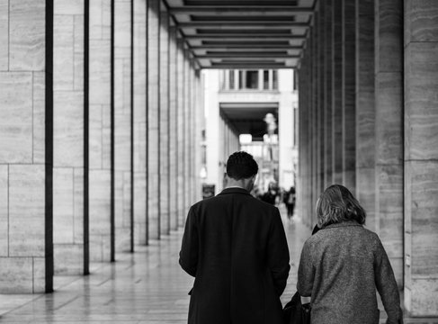 An adult couple walk together though a classical European city. The picture is black and white