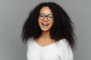Candid shot of optimistic curly woman laughs positively, being in good mood, has healthy skin, looks through transparent glasses, wears white sweater, isolated over grey background. Happiness