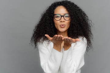 Lovely curly woman sends air kiss, has lips folded, keeps palms outstretched, wears transparent...