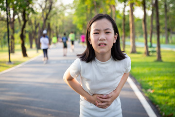 Asian little cute girl holding her hands on belly,suffers from stomach pain,colic after running,exercise in park,sick female child suffering from abdominal pain,period cramps,stomachache concept