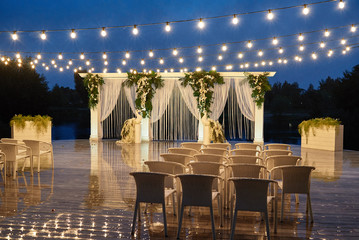 Night wedding ceremony with arch, orchid flowers, chairs and bulb lights in forest outdoors, copy...