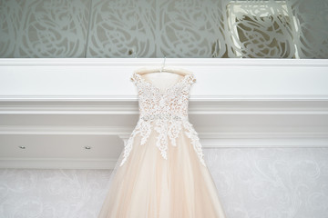 Beautiful beige embroidered wedding dress hanging on hanger against wall in luxury hotel room, copy space, close up. Bridal morning preparations. Wedding concept