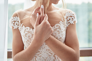Portrait of beautiful bride in wedding dress with crossed hands standing near window in room, copy...