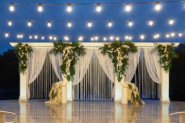 Night wedding ceremony with arch, orchid flowers, palm leaves, chairs and bulb lights in forest...