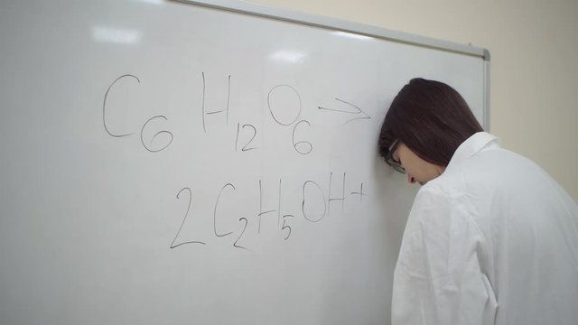 Chemist student in white lab coat trying to write simplified formula for the synthesis of ethanol from glucose, but forgot from frustration banging his head on the board.