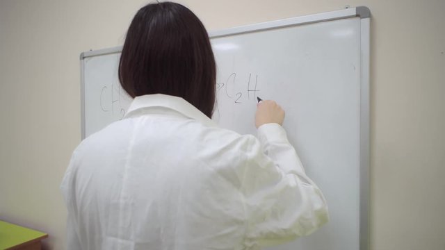 Chemist student writes with errors the general chemical formula for hydrating ethylene with a black marker on blackboard.Girl in white bathrobe is preparing for laboratory work on ethanol.