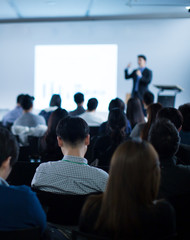 "Speaker giving a talk at a corporate business conference. Audience in hall with presenter in front of presentation screen. Blurred executive giving speech during business and entrepreneur seminar. "