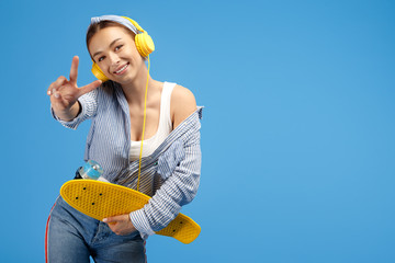 Photo of cheerful young woman with yellow headphones posing with penny or skateboard and showing peace gesture over blue background.