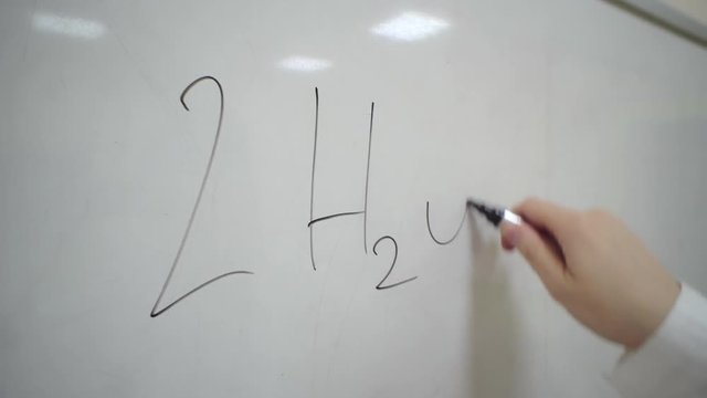 Close-up hand of chemical technician with black marker writes the general formula of water electrolysis on whiteboard.