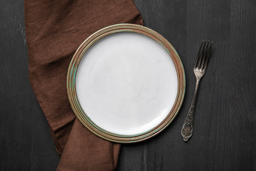 top view of vintage white empty round plate on black wooden table near brown napkin and silver fork