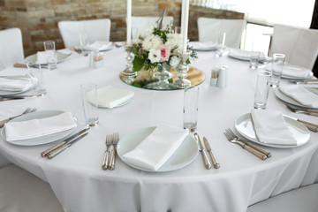 Wedding table setting decorated with fresh flowers. White plates, silverware, white tablecloth and white room. Wedding floristry. Banquet table for guests. Bouquet with roses, hydrangea and eustoma